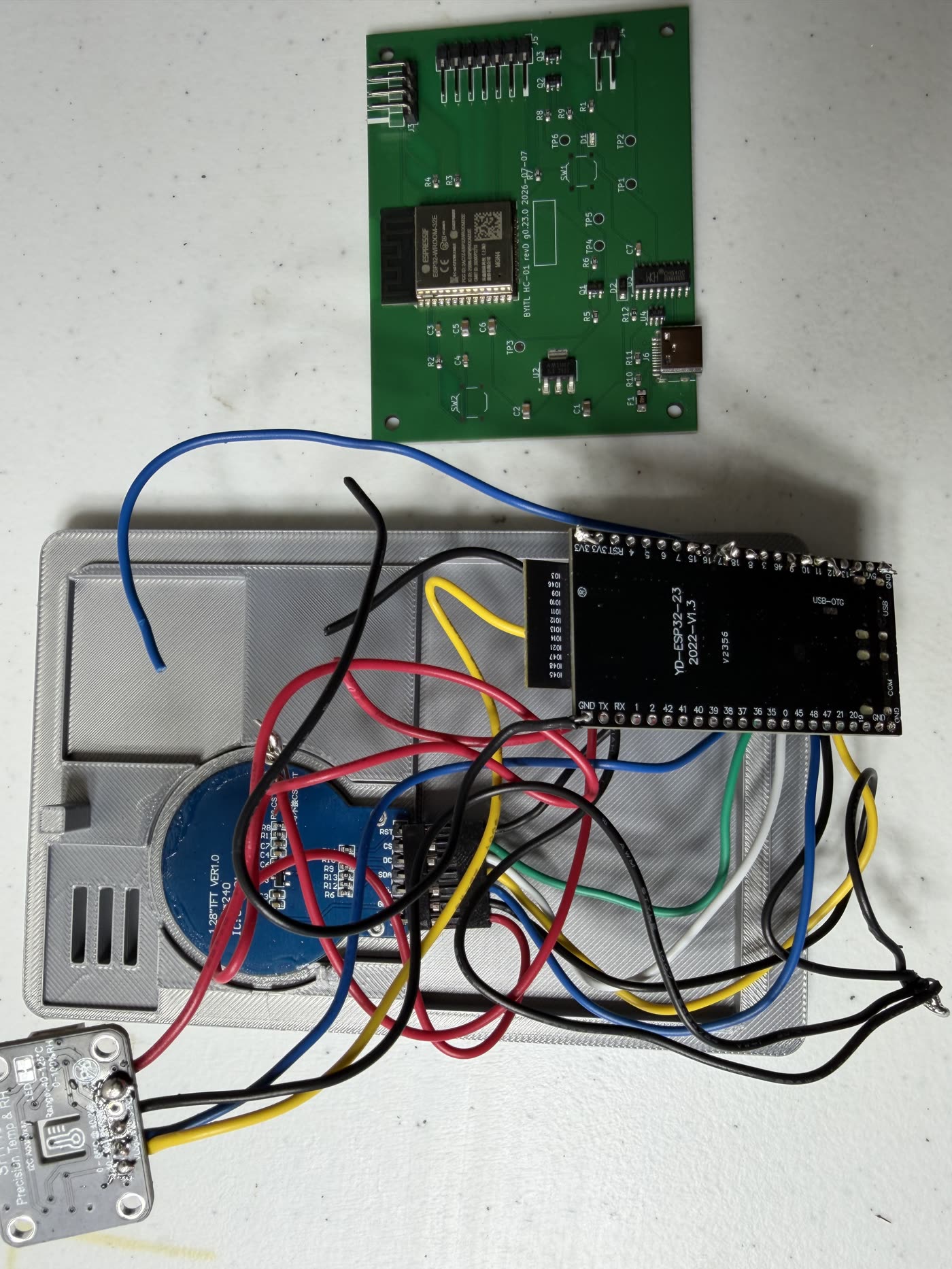 A bare green HC-01 rev D circuit board above the jumper-wire prototype it replaced, which sits on a grey 3D-printed enclosure surrounded by loose wires