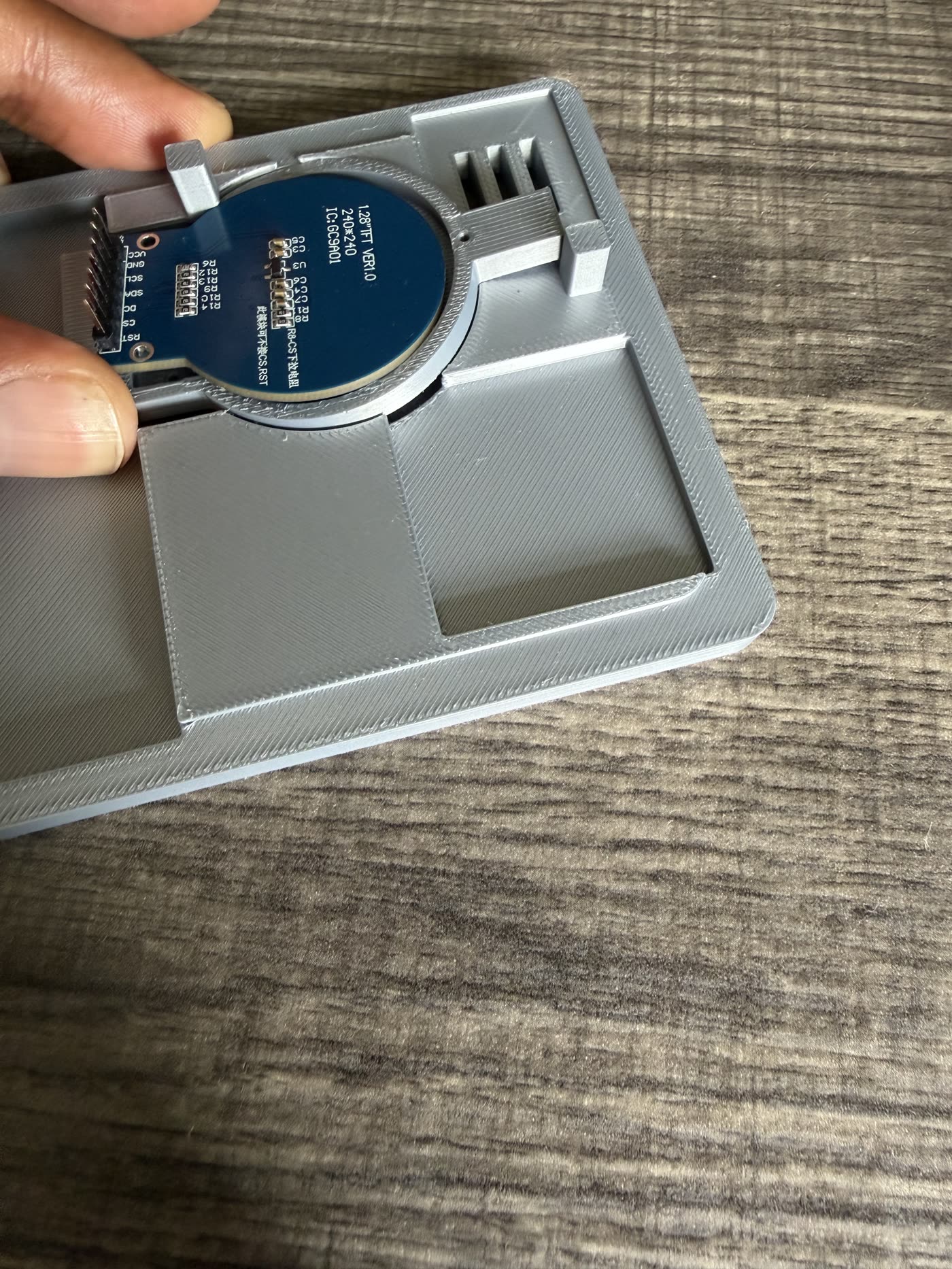 A round 240x240 TFT display module held against a grey 3D-printed enclosure lid with a circular recess and a rectangular cutout